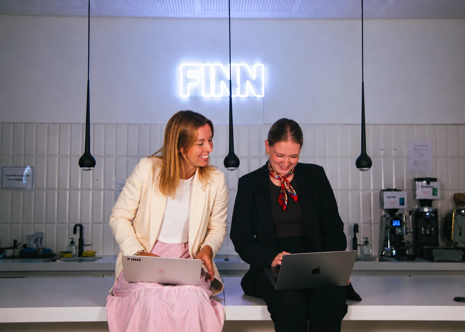 Two FINN colleagues working on laptops beneath the FINN sign in the office kitchen